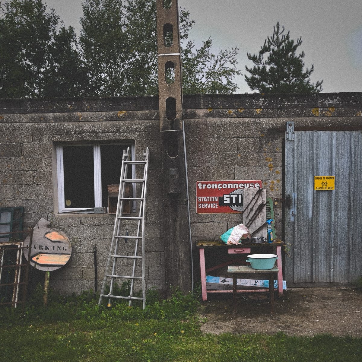 Mur d’atelier à Saint-Ivy, poteau électrique en béton, échelle, panneau tronçonneuse et objets épars.